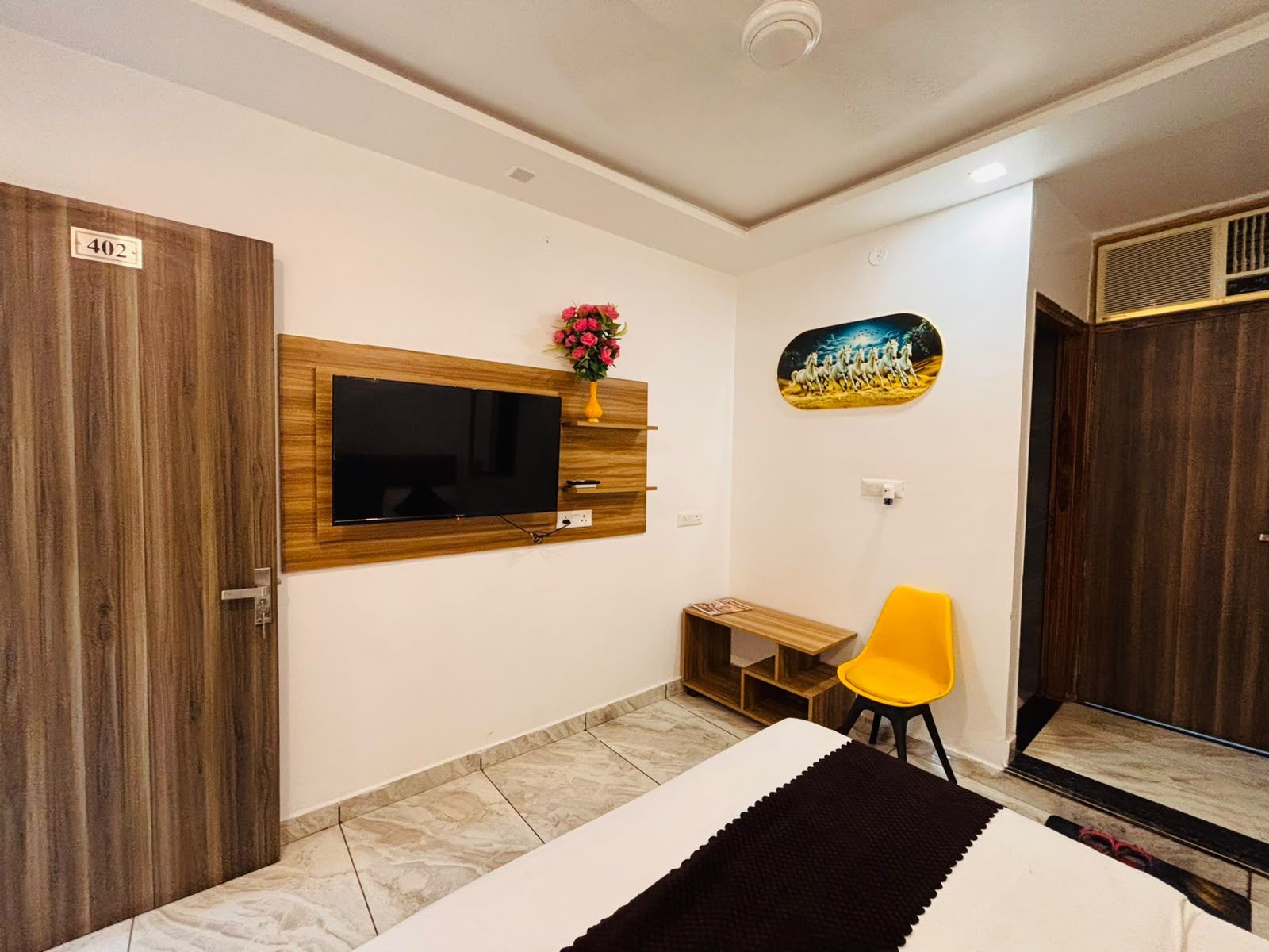 Quarto de hotel com porta de madeira, TV de tela plana na parede, escrivaninha de madeira pequena, cadeira amarela e cama com manta preta.