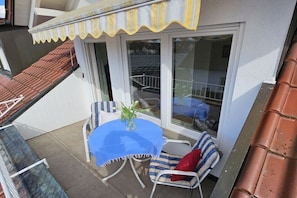 Ein Balkon mit Tisch und Stühlen, einer Topfpflanze und Blick ins Innere durch eine Glastür.