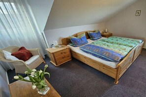 Een slaapkamer met een houten bed, een stoel, een nachtkastje en een vaas met bloemen.