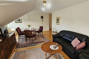 Ein Wohnzimmer mit einem schwarzen Ledersofa, einem Holzes Esstisch und Stühlen. Ein Couchtisch mit Untersetzer steht ebenfalls dort. Eine Uhr und gerahmte Bilder zieren die Wände.