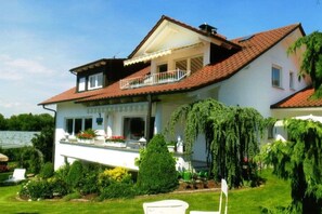 Ein zweistöckiges Haus mit einem Balkon, umgeben von gepflegten Gärten und einem wolkenlosen Himmel.