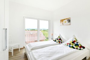 Een slaapkamer met twee bedden, een witte stoel en een raam met uitzicht op een gebouw en groen.