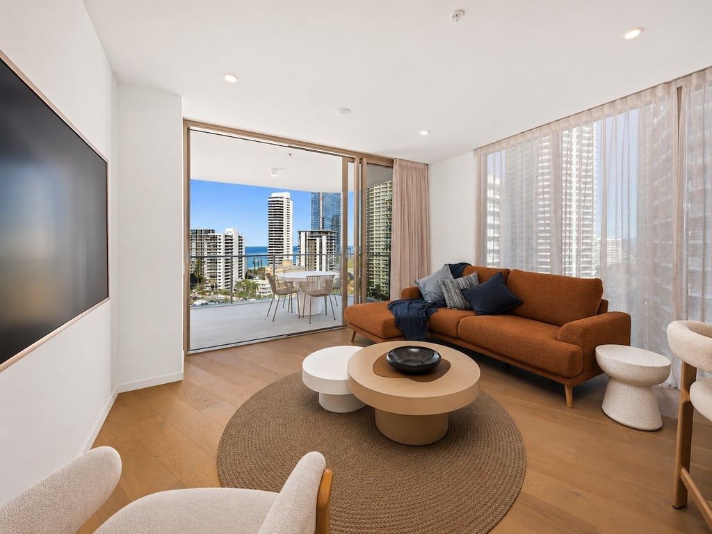 A modern living room with a sofa, coffee table, and chairs, a large TV, and a balcony with a city view.