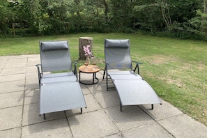 Two lounge chairs with extended footrests on a paved patio, a small round table with a vase of flowers, and a tree stump in the background. - Holiday apartment with 3 bedrooms (Ballum (watteninseln))