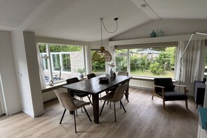 Un comedor con una mesa y sillas de madera oscura, ventanas amplias con vista a zonas verdes y una lámpara colgante.