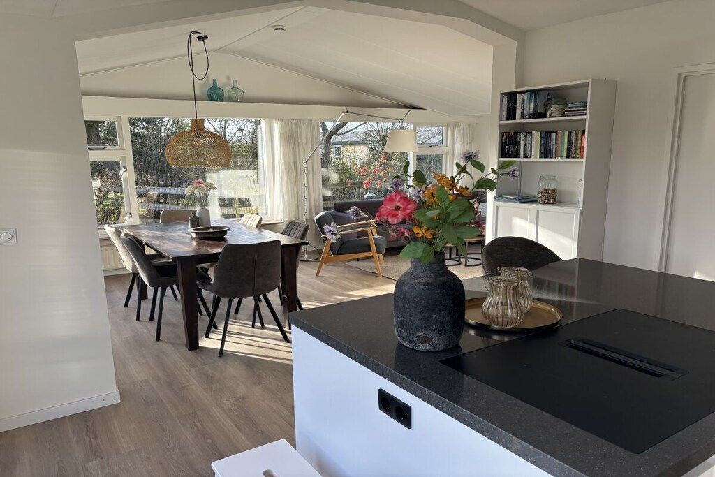 A modern kitchen with a central island, dining area with chairs, and a bookshelf.