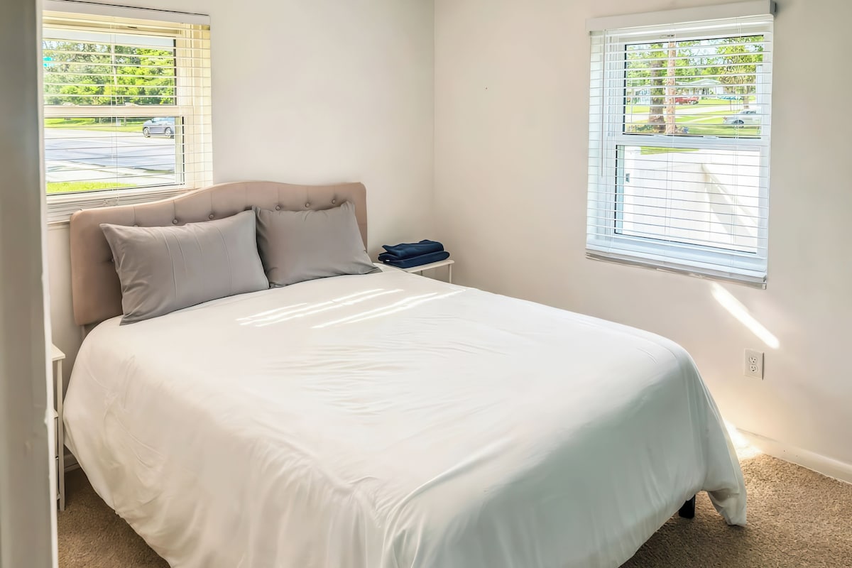 Un dormitorio con cama, dos ventanas y una mesita de noche.