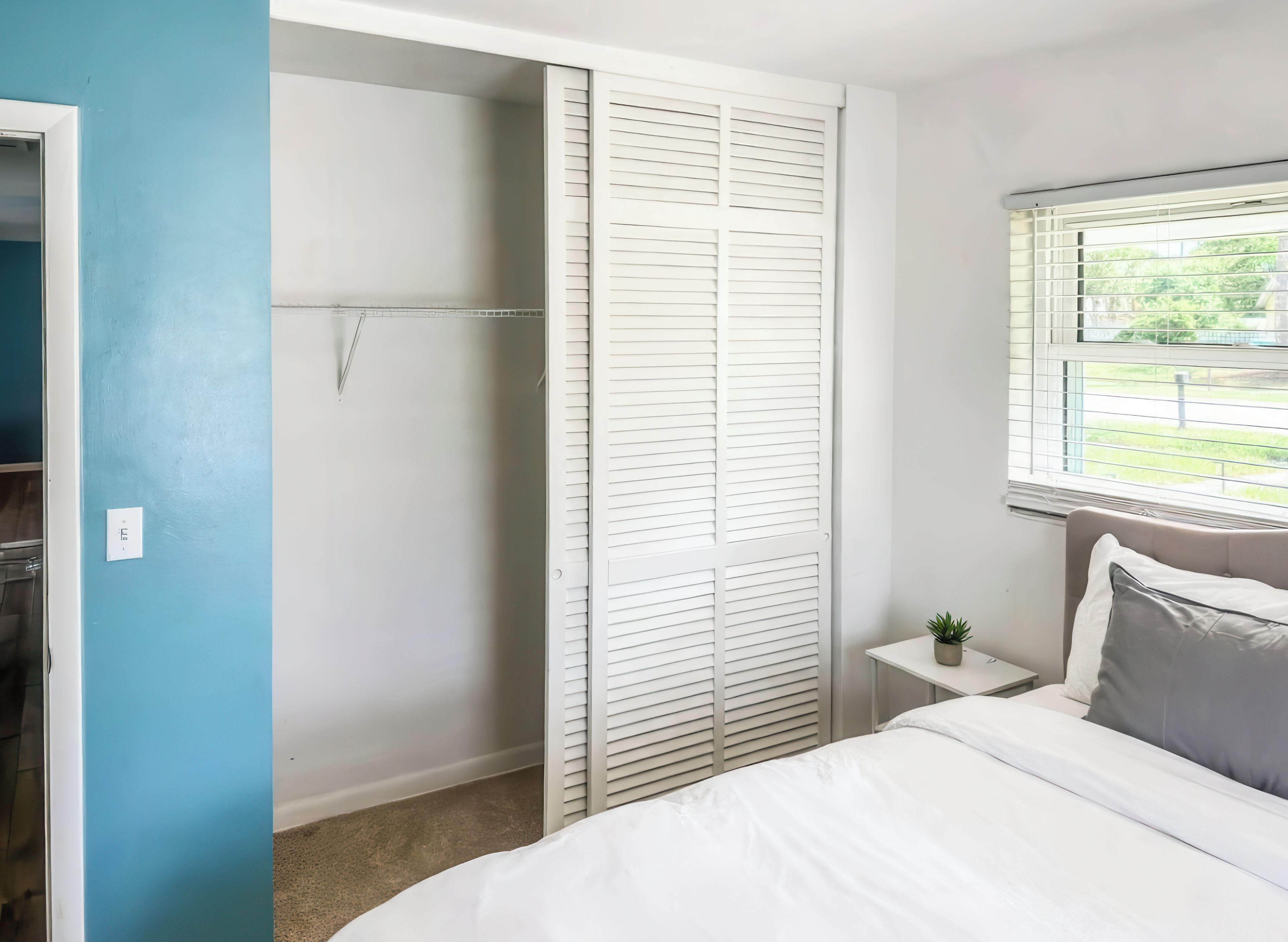 A bedroom with a bed, a nightstand, a window with blinds, and a closet with white shutters.