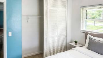 A bedroom with a bed, a nightstand, a window with blinds, and a closet with white shutters.