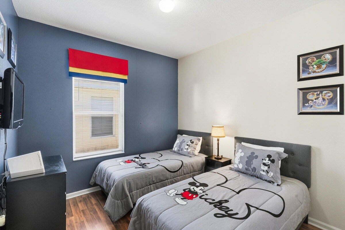 A bedroom with two beds, each with Mickey Mouse-themed bedding, a television, a desk, and a window with blinds.