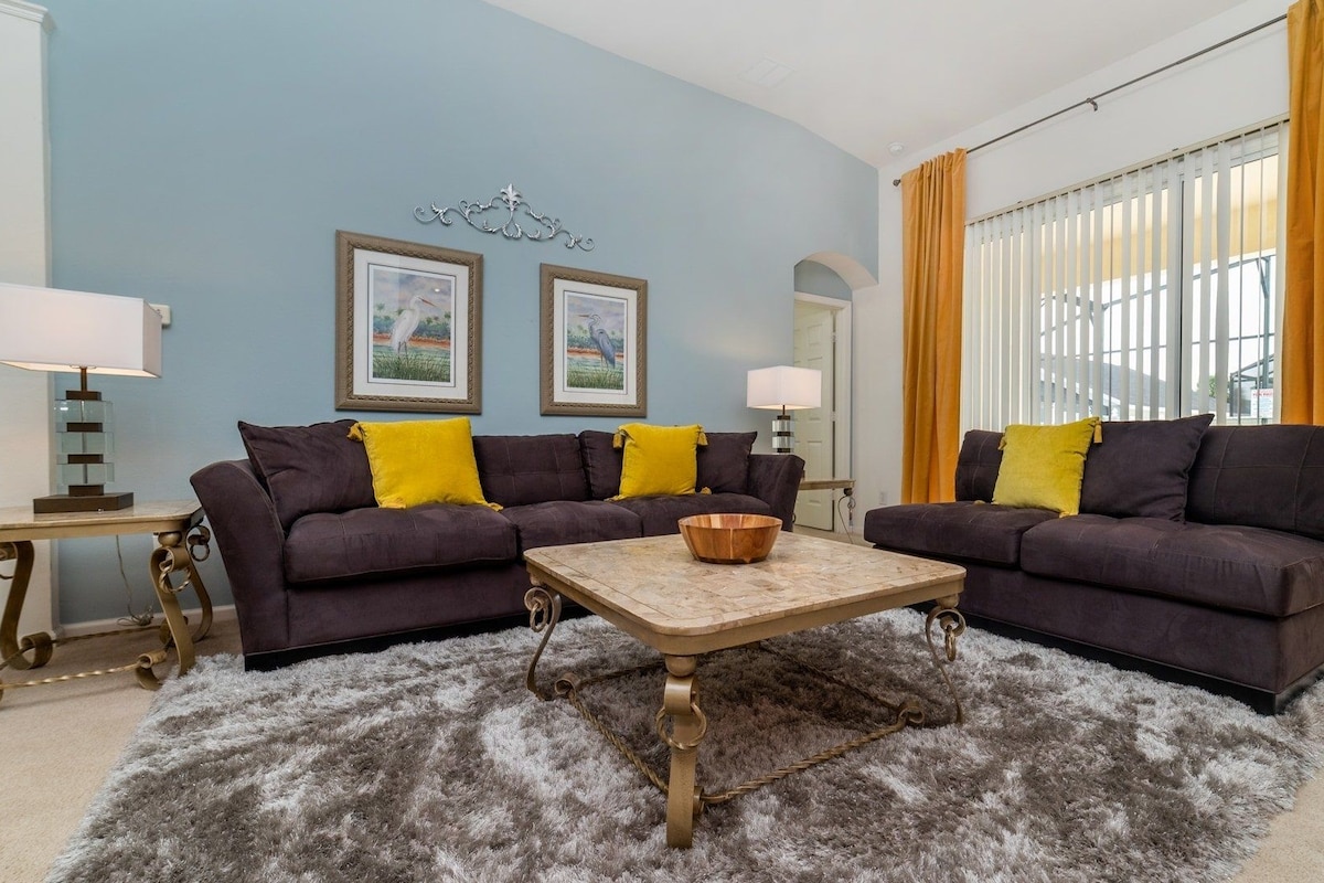 A living room with two dark sofas, a wooden coffee table, and a fluffy rug. There are two framed pictures on the wall, a lamp on a side table, and a window with yellow curtains.