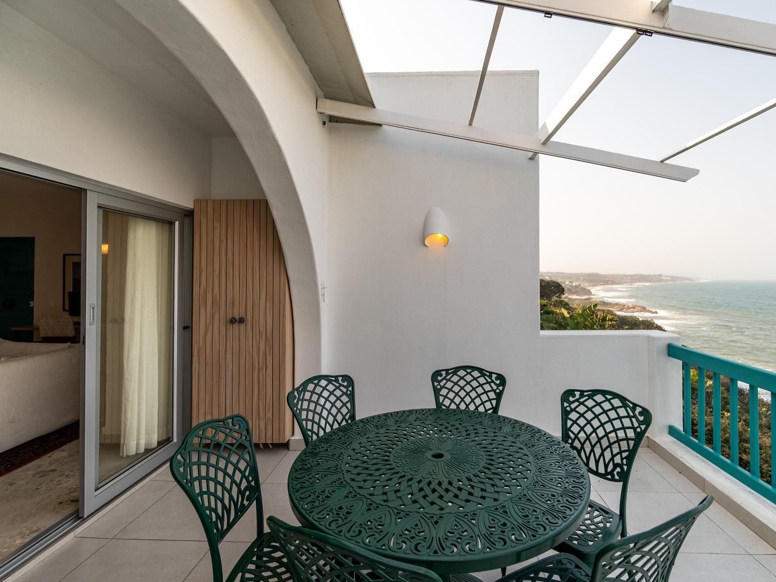 En balkon med et rundt bord og stole, udsigt til havet og en hvid væg med en lysarmatur.