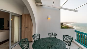 En balkon med et rundt bord og stole, udsigt til havet og en hvid væg med en lysarmatur.
