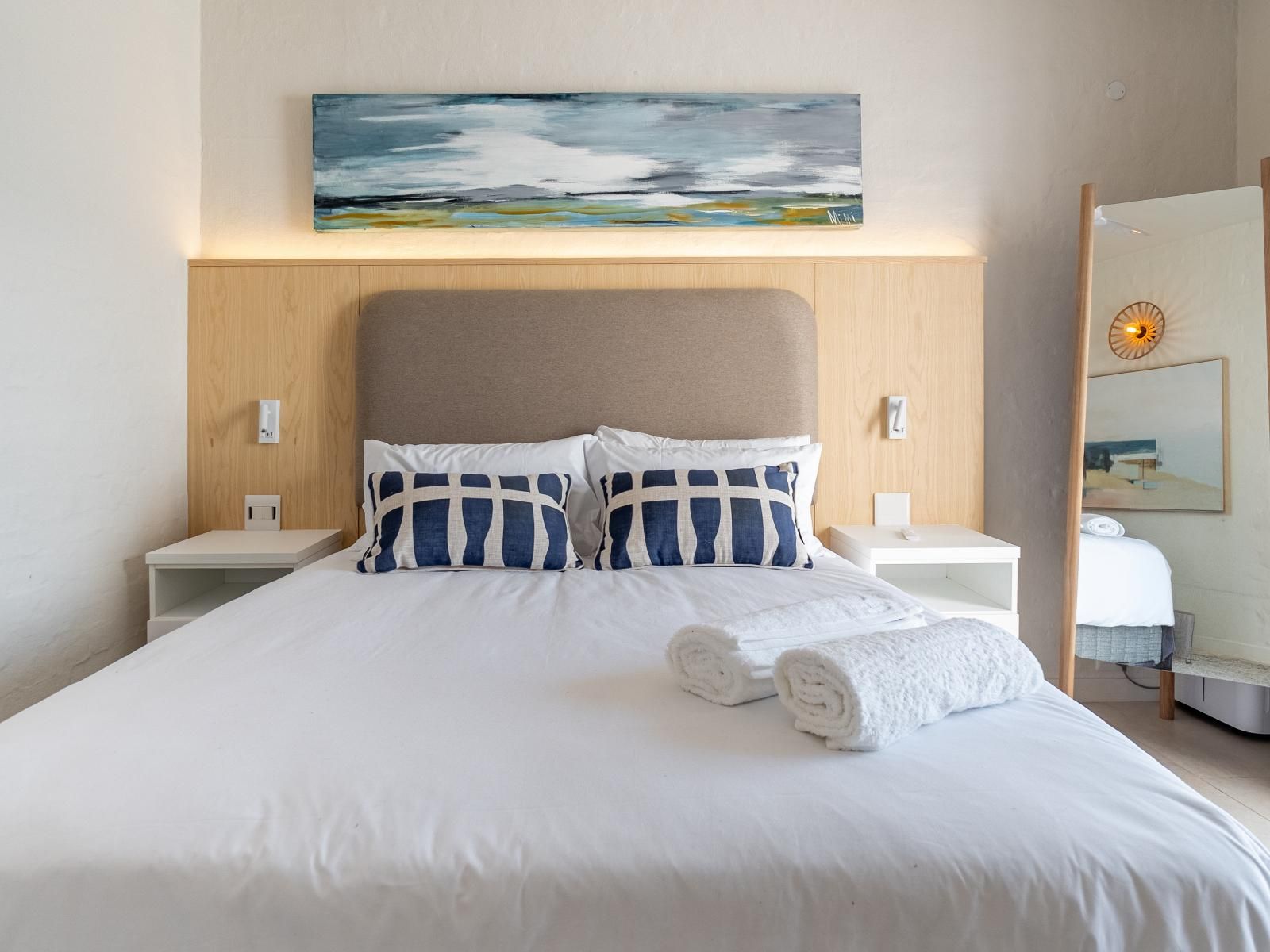 En pænt redt seng med hvide sengetøj og blå puder, et billede over sengegavl og et natbord med en lampe.