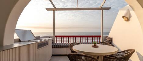 A sunlit outdoor seating area with a round table and wicker chairs, overlooking a body of water.