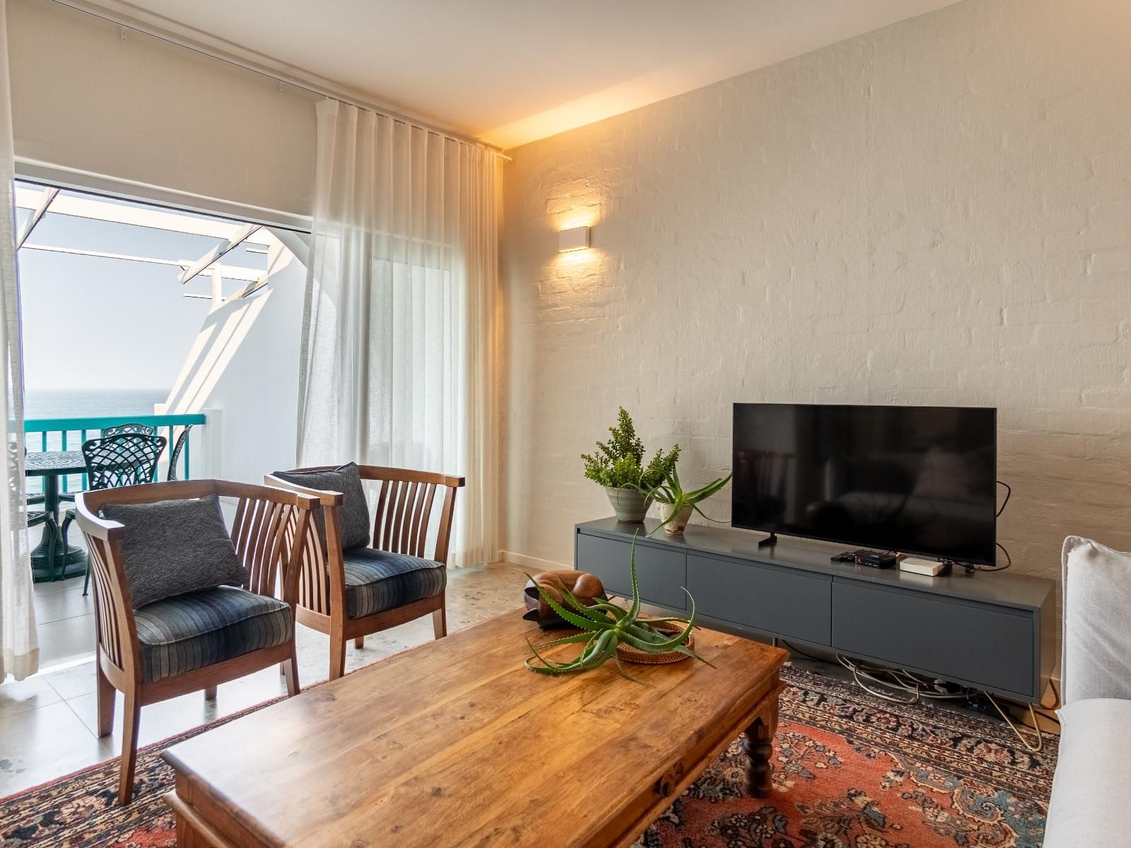 A hotel room with a wooden dining table, two chairs, a television on a grey cabinet, and a view of the sea through a balcony.