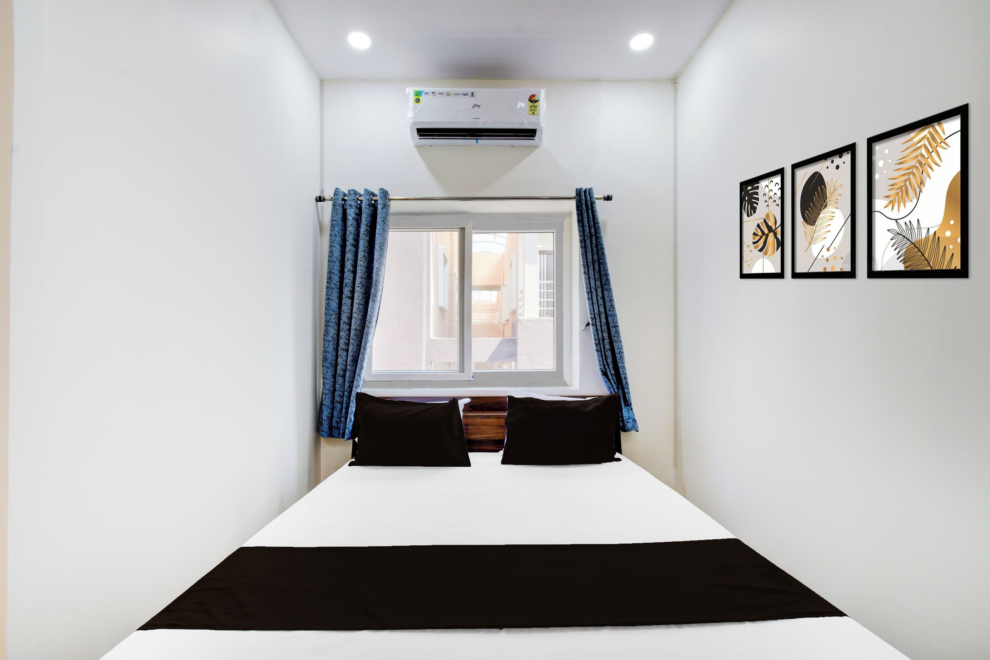 A hotel room with a bed, curtains, an air conditioning unit, and framed artwork on the wall.