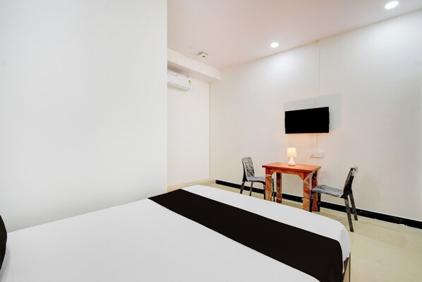 A hotel room with a bed, a desk, two chairs, a television mounted on the wall, and an air conditioning unit.