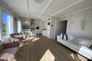A modern living room with a white sofa, a dining table, and chairs. There is a kitchen area with white cabinets and a window. - Type B + "Zee, Bos en Duin" (Zoutelande)