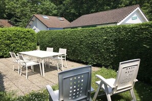 A patio with a table and chairs, a hedge, and a building with a tiled roof in the background. - Type B + "Zee, Bos en Duin" (Zoutelande)