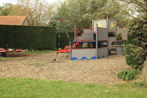 A playground with a slide and climbing structures, surrounded by a hedge and a building with a tiled roof. - Type B "Zee, Bos en Duin" (Zoutelande)