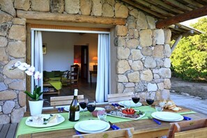 An outdoor dining area with stone walls, a wooden table set for a meal, and a view into a room with a sofa and lamp. - Holiday home with 2 bedrooms (Porto Vecchio)