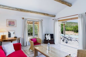 Ein Zimmer mit einem roten Sofa, einem Holztisch, einem Korbstuhl, einem Fernseher und Blick auf einen Pool und Grünflächen.