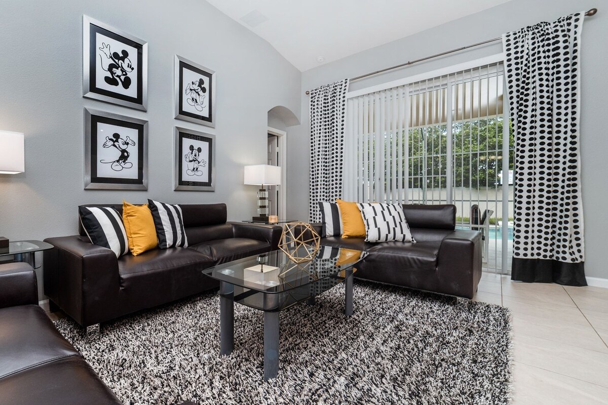 A modern living room with two black leather sofas, a glass coffee table, and a black and white patterned rug. There are framed pictures on the wall, a floor lamp, and a window with black and white curtains.