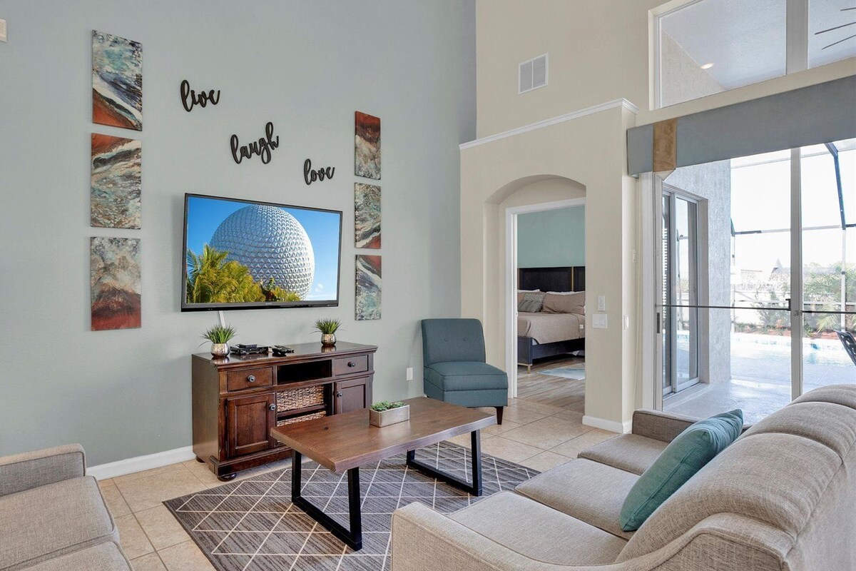 A modern living room with a flat-screen TV, a coffee table, and a sofa. There are wall art pieces and a sign that says "live, laugh, love."