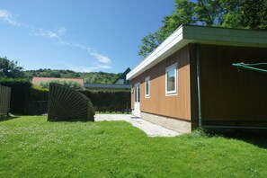 A house with a wooden exterior, a visible window, and a garden area with grass and a hedge. - Holiday apartment with 1 bedroom (Zoutelande)