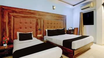 A hotel room with two beds, wooden headboards, a nightstand with a lamp, a small table with a vase of flowers, and an air conditioning unit.