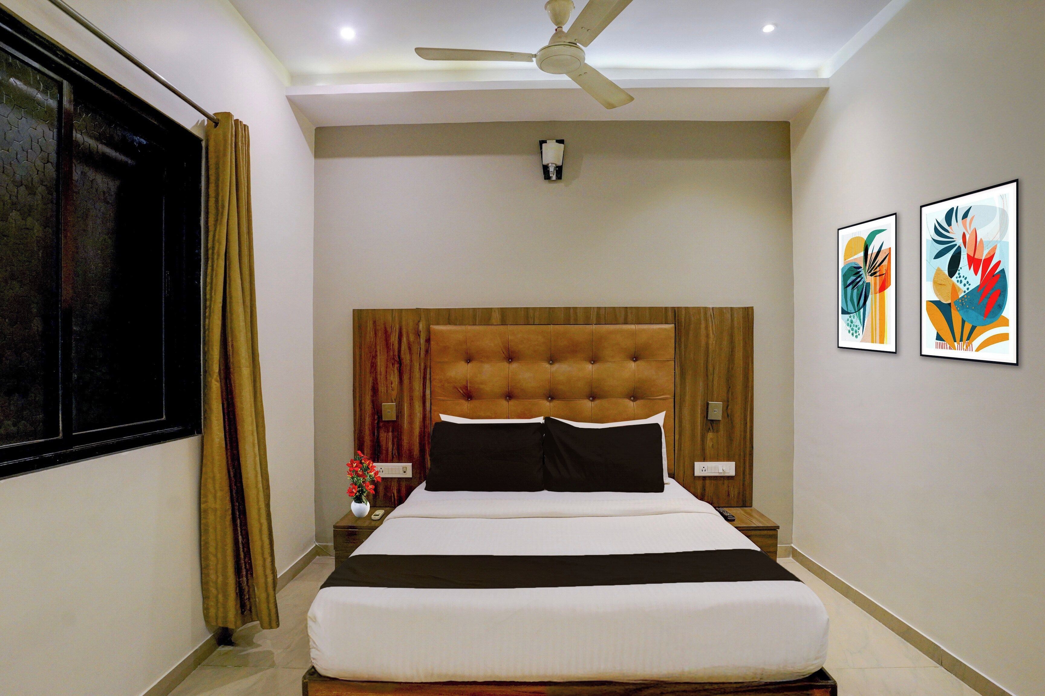 A hotel room with a bed, a ceiling fan, a nightstand, a vase with flowers, and two framed artworks on the wall.