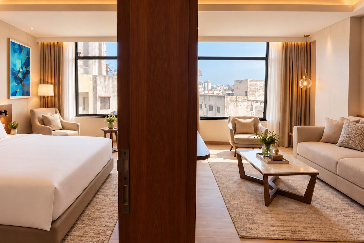 A modern hotel room with a large bed, two armchairs, a sofa, a coffee table, and a view of the cityscape through large windows.