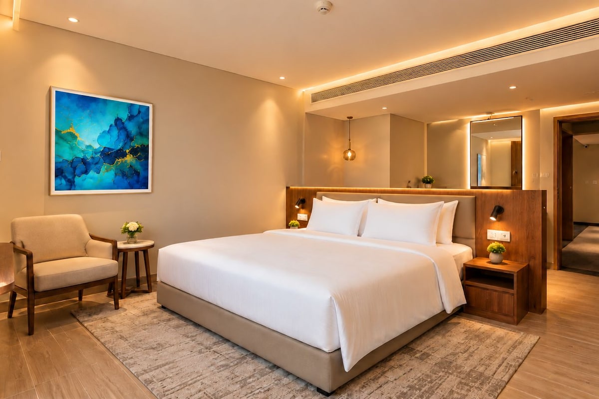 A hotel room with a large bed, a chair, a small table with a vase, a painting on the wall, and a mirror.