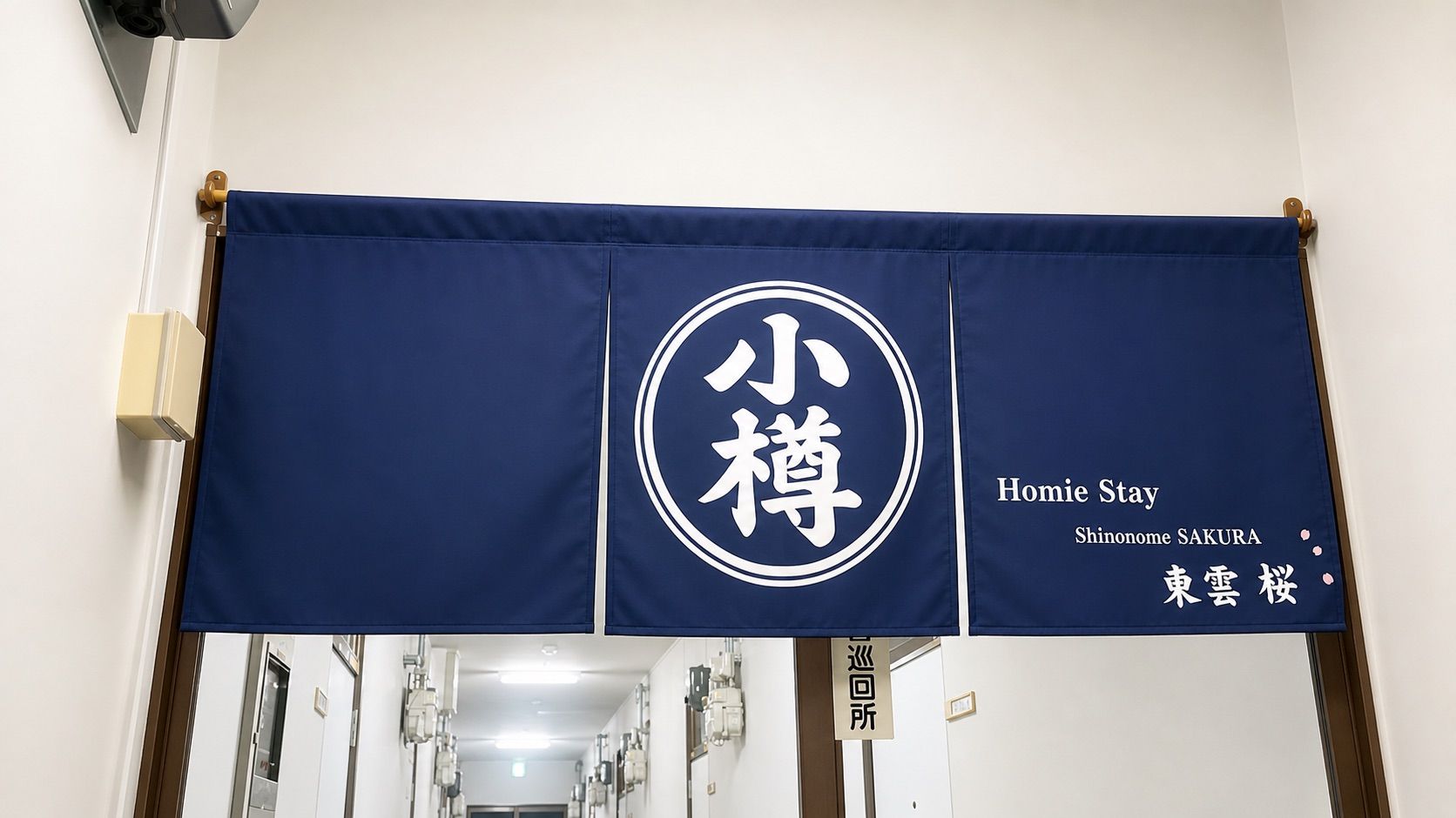 Een banner met Japanse tekens en Engelse tekst, waarop een hotel genaamd 'Homie Stay Shinonome SAKURA' staat, met een bordje voor de receptie.
