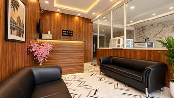 A hotel reception area with a wooden reception desk, a black leather sofa, a potted plant, and a check-in/out sign.