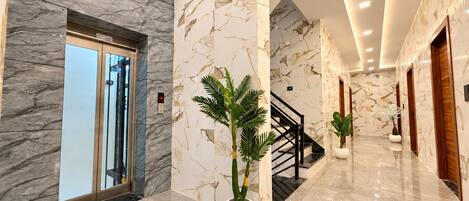 Un moderno vestíbulo de hotel con paredes de mármol, un ascensor de vidrio, una escalera y plantas en macetas.