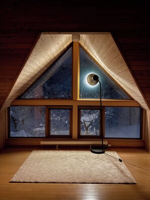 Ein Zimmer mit schrägen Wänden, großen Fenstern und einer Stehlampe.