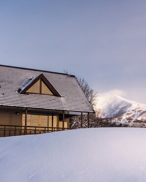 A building with a sloped roof, large windows, and a view of a snow-covered landscape with mountains in the background. - Mountain View Niseko 【Private Cottage with Great Views】 (ニセコ町)