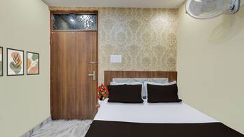 A bedroom with a wooden door, a bed with white and black bedding, a wall-mounted fan, and patterned wallpaper.