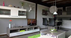 A modern kitchen with a built-in oven, a dining area with a table and chairs, and a living room with a flat-screen TV. - Vacation Rental Between Chiemsee and Salzburg in the Pre-Alps *Wi-Fi, Thermomix, Balcony, View* (Siegsdorf)