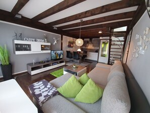 Uma sala de estar moderna com um sofá cinzento, almofadas verdes, uma TV de tela plana e um espaço de jantar com uma mesa e cadeiras de madeira.