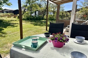 Une terrasse extérieure avec une table dressée pour deux personnes, agrémentée d’un plateau vert sur lequel sont posés deux mugs, un pot de fleurs rose et un bol.