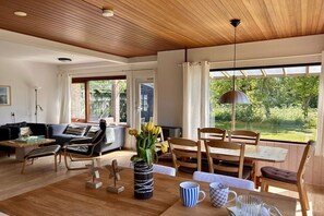 A modern living room with a wooden ceiling, a sofa, a dining table with chairs, and a view of greenery outside. - Bungalow with 3 bedrooms (Julianadorp)