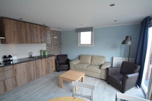 Un espacio habitable moderno con cocina, un sofá color beige, dos sillones, una mesa de centro de madera y una ventana con cortinas.