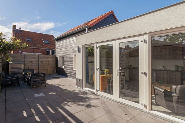 Un patio moderno con puerta corrediza de vidrio que da a una cocina, cerca viva de madera y mobiliario exterior.