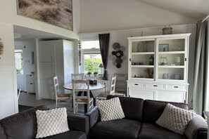A modern living room with a dark leather sofa, white cushions, a white cabinet with glass doors, and a dining area with a round table and chairs. - Holiday home with 3 bedrooms (Julianadorp)