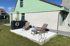 A house with a green exterior, a porch with outdoor furniture, and a brick wall. - Holiday home with 3 bedrooms (Julianadorp)
