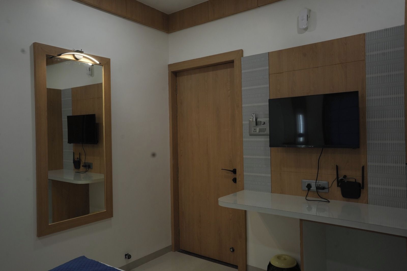 Een badkamer met een houten deur, een spiegel met verlichting, een flatscreen televisie en een witte wastafel.
