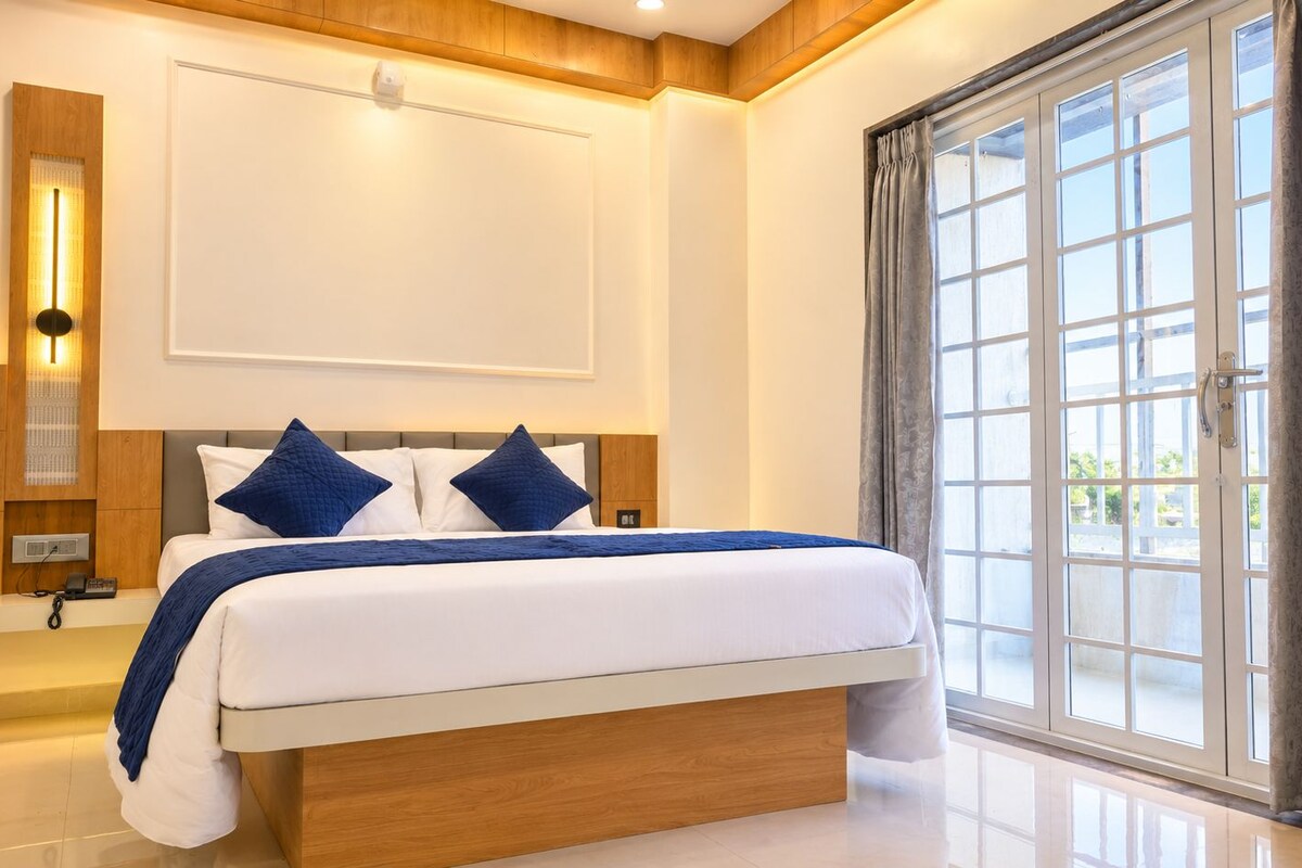 A modern bedroom with a large bed, a nightstand, a wall-mounted light, and a view through a large window.